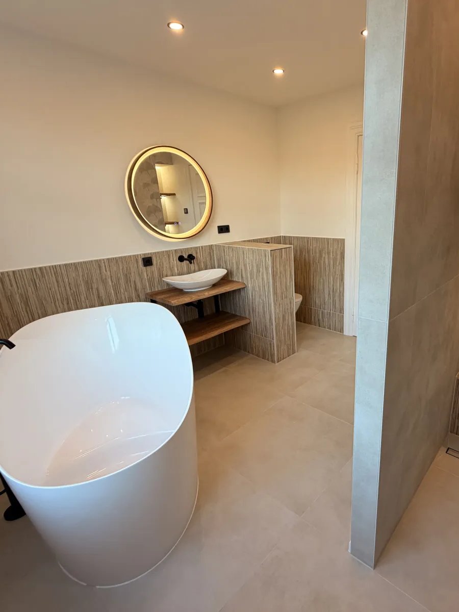 Fertiges Bad: freistehende Wanne, runder beleuchteter Spiegel über dem Aufsatzbecken und Dusche hinter einer gefliesten Trennwand