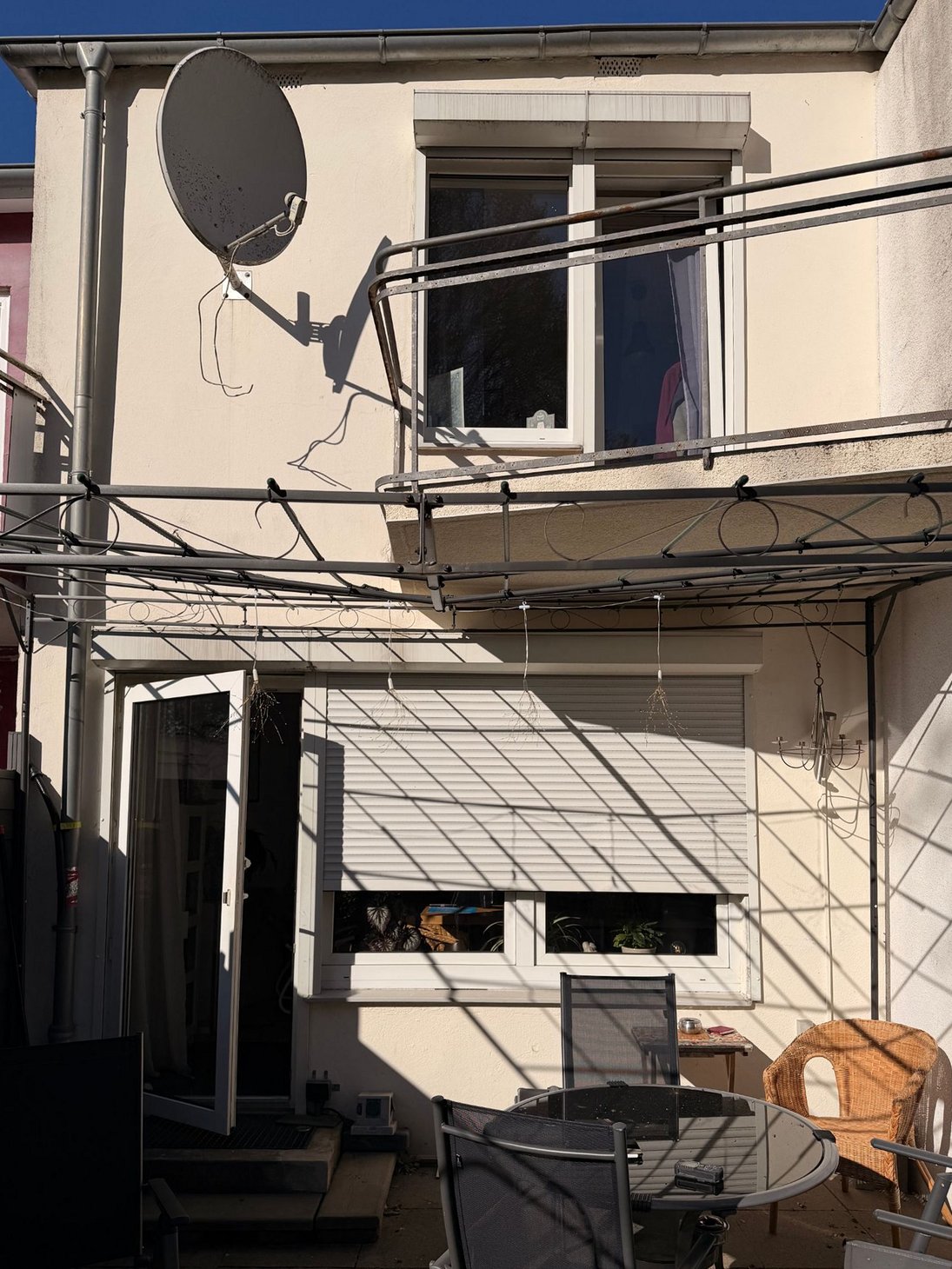 Gartenseite vorher: hell verputzte Fassade mit Satellitenschüssel, Balkon mit Metallgeländer, weißen Fenstern und Pergola über der Terrasse