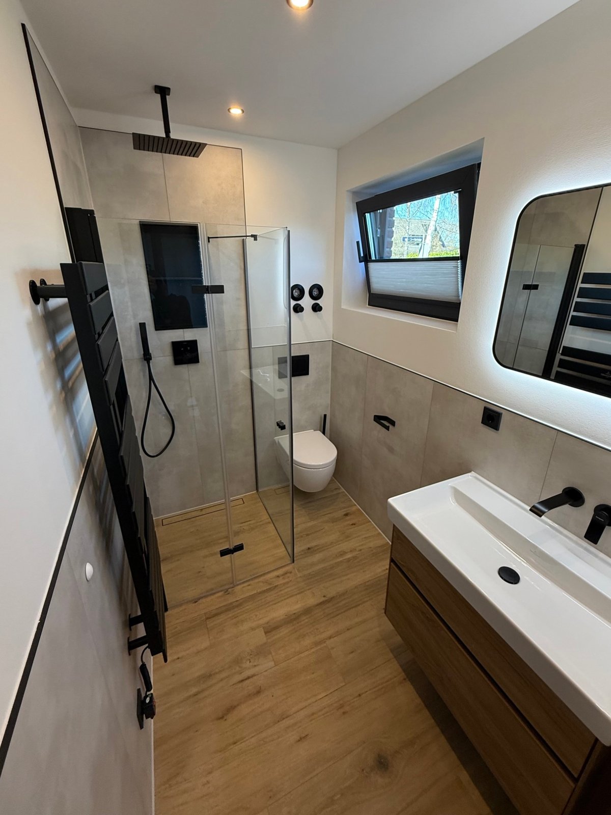 Gästebad nachher: bodengleiche Dusche mit Glaswand und schwarzer Regendusche, Wand-WC, großformatige Fliesen in Betonoptik, Boden in Holzoptik, Waschtisch mit Holzunterschrank und beleuchteter Spiegel